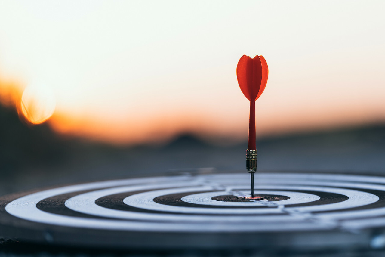 Close up red dart arrow hitting target center dartboard on sunset background. Business targeting and focus concept